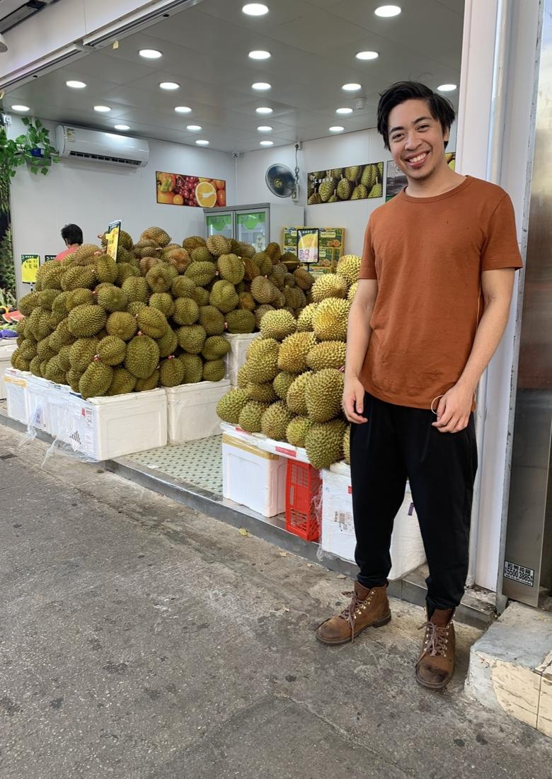 Michael smiling next to piles of durian at a fruit shop in Hong Kong